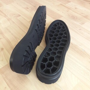 K&ouml;nnte beinhalten: Zwei schwarze Schuhsohlen werden auf einer hellbraunen Holzoberfl&auml;che gezeigt. Eine Sohle hat ein Profilmuster, die andere ein Wabenmuster. Die Sohlen bestehen aus einem strapazierf&auml;higen Material.