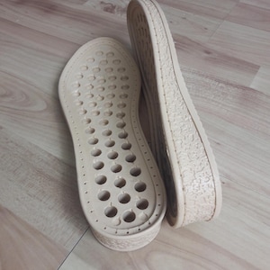 Puede incluir: Dos suelas de zapatos beige con un patrón texturizado en la parte inferior. Una suela tiene una fila de agujeros a lo largo del lado.