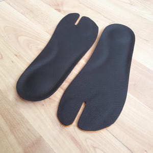 Puede incluir: Un par de plantillas negras texturizadas diseñadas con una separación para los dedos. Las plantillas tienen una forma contorneada para apoyar el pie, con una separación distintiva entre el dedo gordo y los demás, y una base fina de color.