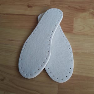 Puede incluir: Dos plantillas de fieltro blanco con una serie de pequeños agujeros espaciados uniformemente a lo largo de los bordes. Las plantillas, de forma ovalada, están diseñadas para colocarse dentro de los zapatos. Se encuentran sobre una superficie de madera marrón claro.