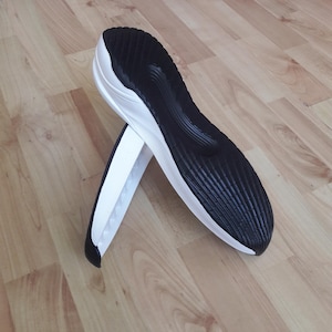 Puede incluir: Dos suelas de zapatillas deportivas, una negra y otra blanca, están cruzadas. La suela negra tiene un patrón texturizado y acanalado en la parte inferior. La suela blanca tiene un diseño liso y curvado. Los zapatos están sobre un suelo de madera.