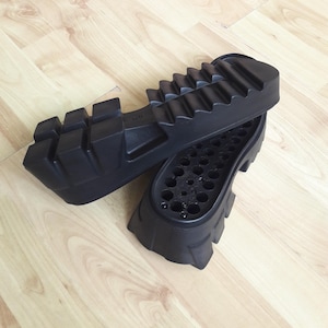 May include: Two black rubber shoe soles with a chunky, ridged design. One sole is positioned above the other, revealing the interior with multiple small holes. The soles are likely for boots or heavy-duty footwear.