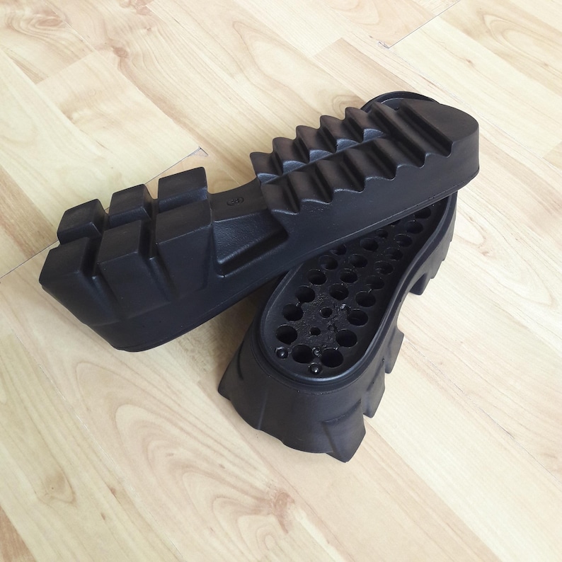 May include: Two black rubber shoe soles with a chunky, ridged design. One sole is positioned above the other, revealing the interior with multiple small holes. The soles are likely for boots or heavy-duty footwear.