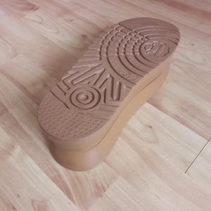 Könnte beinhalten: Eine hellbraune Plateausohle mit einem strukturierten Profilmuster. Die Sohle ist dick und hat eine abgerundete Spitze und Ferse. Die Schuhsohle liegt auf einem Holzboden.