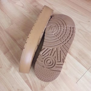 May include: Two tan-colored shoe soles are displayed. One sole is upright, showing the side profile with a ridged design. The other sole is lying flat, revealing a textured pattern with a circular design and the number "10".