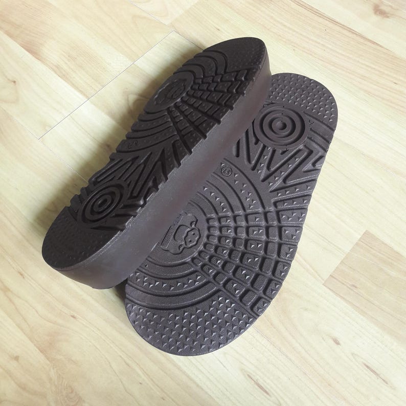 May include: Two brown shoe soles with textured patterns and a raised heel. The soles are made of a dark brown material and feature various geometric designs, including circles and lines. The soles are positioned on a light-colored wooden surface.