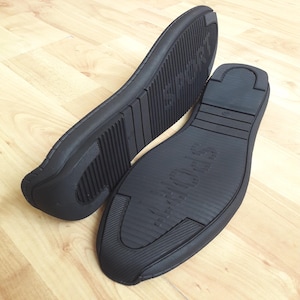 May include: Two black shoe soles with the word "SPORT" embossed on the bottom. The soles have a textured, grooved design for traction. One sole is positioned at an angle, while the other is flat, both on a wooden surface.