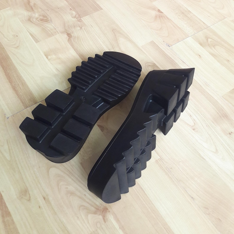 May include: Two black rubber shoe soles with a chunky, ridged design. The soles are new and appear to be for boots or heavy-duty footwear. The soles are lying on a light-coloured wooden surface.