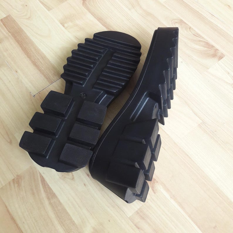 May include: Two black rubber boot soles with a deep tread pattern are shown on a light wood surface. The soles are thick and have a rugged design, suitable for outdoor footwear. The image highlights the texture and construction of the soles.