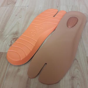 May include: Two split-toe shoe insoles. One is orange with a textured surface and the other is tan. The insoles are designed to fit shoes with a separate compartment for the big toe.