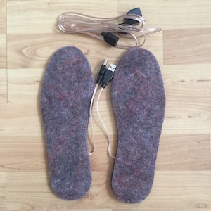 K&ouml;nnte beinhalten: Ein Paar graue Heizsohlen mit USB-Kabeln. Die Sohlen bestehen aus einem weichen, flauschigen Material.