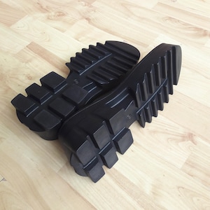 May include: A pair of black boot soles with a deep tread pattern are shown on a light wood-grain surface. The soles are thick and have a rugged, industrial design, suitable for creating durable footwear.