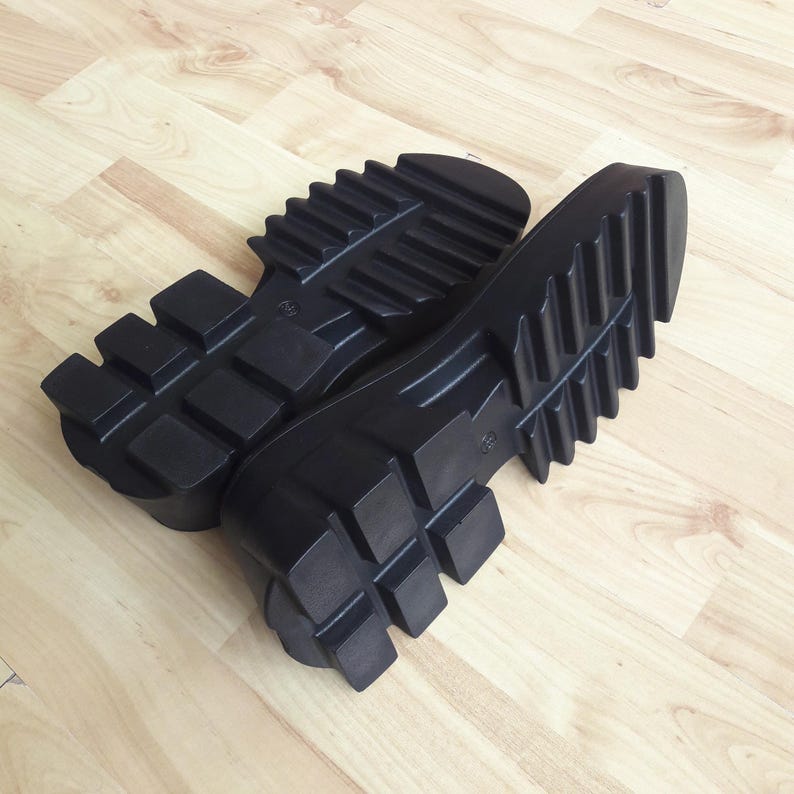 May include: A pair of black boot soles with a deep tread pattern are shown on a light wood-grain surface. The soles are thick and have a rugged, industrial design, suitable for creating durable footwear.