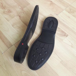 Puede incluir: Dos suelas de zapatos negras con patrones texturizados y un punto rojo en una de ellas. Las suelas están hechas de un material duradero y están diseñadas para calzado. La imagen muestra la parte inferior de las suelas, revelando la banda de rodadura.