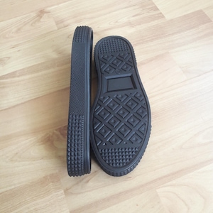 Puede incluir: Una suela de zapato de goma negra con un lateral texturizado y una banda de rodadura con patrón de diamantes en la parte inferior. La suela tiene una construcción gruesa y duradera con un diseño geométrico y una pequeña muesca rectangular.