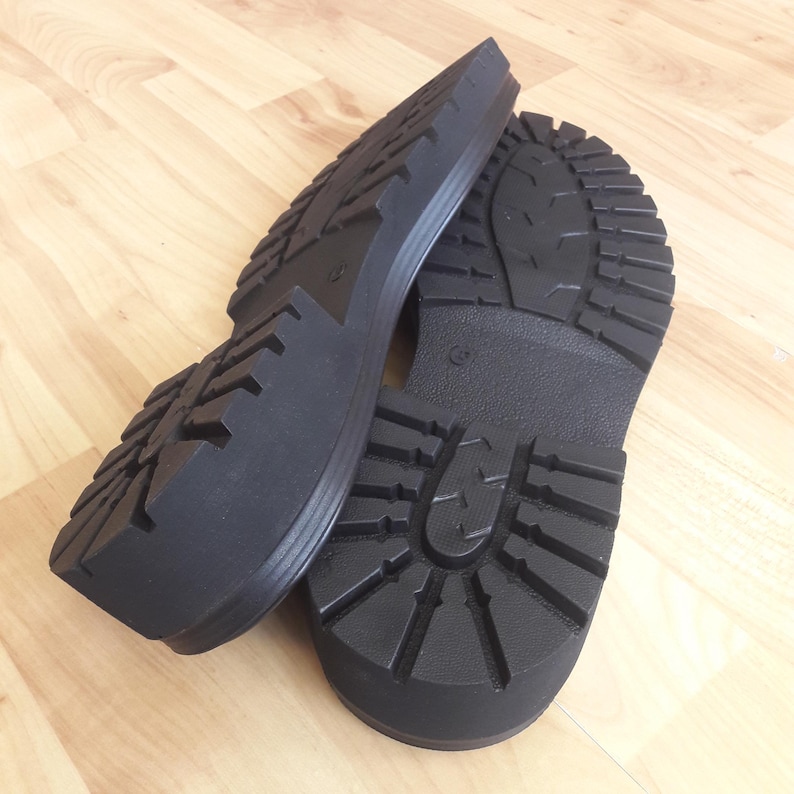 K&ouml;nnte beinhalten: Zwei schwarze Gummistiefelsohlen mit tiefem Profilmuster werden auf einer hellen Holzoberfl&auml;che gezeigt. Die Sohlen sind dick und haben ein robustes Design, das f&uuml;r Outdoor-Aktivit&auml;ten geeignet ist.