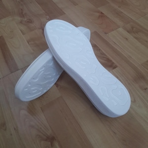 May include: Two white shoe insoles with a textured pattern are displayed on a wooden surface. The insoles are oval-shaped and have a raised, swirling design on the bottom. The insoles are clean and appear new.