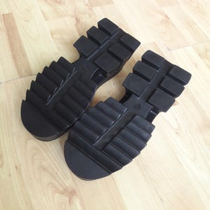 May include: Two black shoe soles with a chunky, ridged tread. The soles have a thick platform and are designed for robust footwear. The soles are made of a durable material, suitable for various shoe styles.