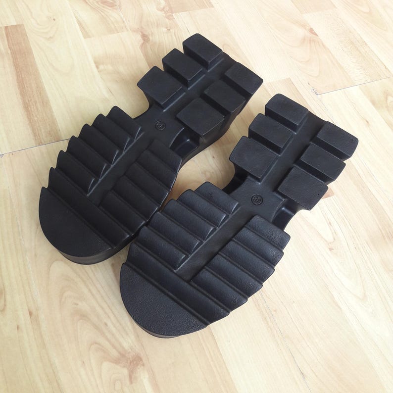 May include: Two black shoe soles with a chunky, ridged tread. The soles have a thick platform and are designed for robust footwear. The soles are made of a durable material, suitable for various shoe styles.