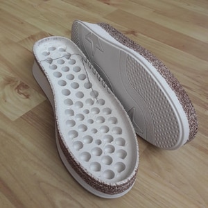 May include: Two white shoe insoles with a perforated design and a brown glittery border. The soles are on a wooden surface. One insole is positioned in front of the other.
