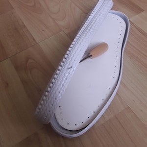 Op de afbeelding: Witte schoenzool met een witte rubberen buitenzool en een gereedschap om te naaien. De zool heeft een patroon van kleine gaatjes.