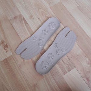 May include: A pair of light gray shoe insoles designed with a split-toe feature. The surface of each insole features a textured pattern and multiple circular areas with small ventilation holes for airflow, providing a distinct shape for footwear.