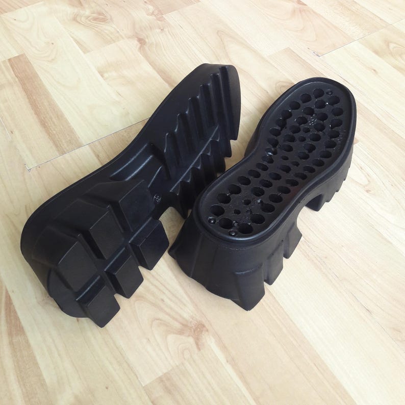 May include: Two black shoe soles with a chunky, lugged design. One sole is upside down, revealing a pattern of small holes. The soles are made of a durable material, suitable for boots or other footwear.