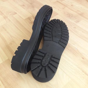May include: Two black rubber boot soles with a lugged tread pattern. One sole is positioned upright, revealing the interior, while the other lies flat, showcasing the tread design. The soles are designed for durability and traction.