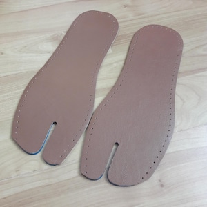 May include: Two tan leather tabi shoe insoles with a split toe design. The insoles have a series of small holes around the edges for stitching. The bottom edge of the split toe is blue.