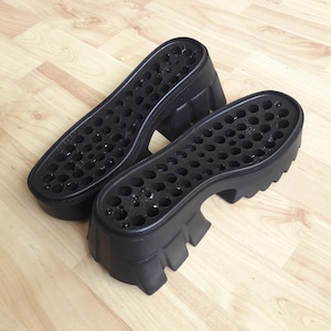 May include: A pair of black platform shoe soles with a chunky heel and numerous small holes on the sole. The soles are made of a solid, matte black material, designed for a bold, edgy look. The soles are approximately 20 cm long.