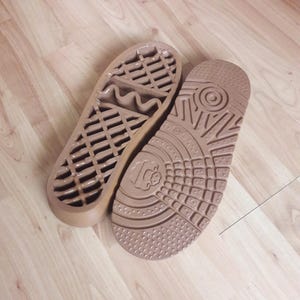 May include: Two tan-colored shoe soles with textured patterns. One sole features a series of parallel lines and a wavy design, while the other has a circular pattern and a small logo. The soles are designed for footwear.