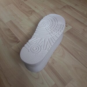 May include: A white platform shoe sole with a textured bottom, featuring a circular design and various patterns. The sole is thick and appears to be made of a durable material. The shoe sole is shown on a wooden surface.