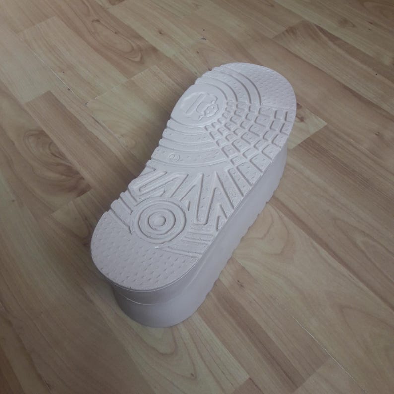 May include: A white platform shoe sole with a textured bottom, featuring a circular design and various patterns. The sole is thick and appears to be made of a durable material. The shoe sole is shown on a wooden surface.