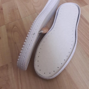 Op de afbeelding: Twee witte schoenzool, een met een getextureerde buitenrand en de andere met een gladde buitenrand en een patroon van kleine gaatjes.