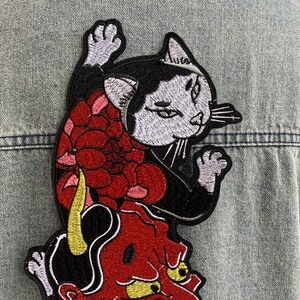 Peut inclure: Un &eacute;cusson brod&eacute; repr&eacute;sentant un chat noir et blanc avec une patte lev&eacute;e, plac&eacute; au-dessus d'un masque de d&eacute;mon rouge aux yeux et &agrave; la corne jaunes, agr&eacute;ment&eacute; d'une grande fleur rouge, le tout cousu sur un tissu en jean bleu clair.