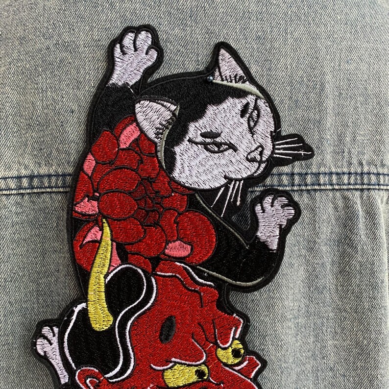 Peut inclure: Un &eacute;cusson brod&eacute; repr&eacute;sentant un chat noir et blanc avec une patte lev&eacute;e, plac&eacute; au-dessus d'un masque de d&eacute;mon rouge aux yeux et &agrave; la corne jaunes, agr&eacute;ment&eacute; d'une grande fleur rouge, le tout cousu sur un tissu en jean bleu clair.
