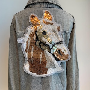 Op de afbeelding: Een spijkerjack met een grote pailletten-paardenkop-patch op de rug. Het paard is bruin, wit en goud.