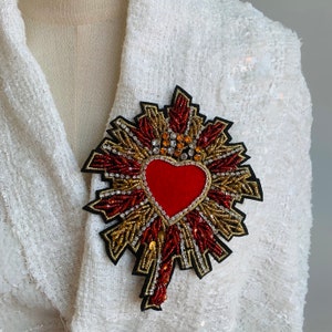 Può includere: Un cuore rosso con decorazioni dorate e rosse, circondato da un'aureola dorata con perline rosse e dorate. Il cuore è centrato al centro del design.