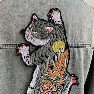 Peut inclure: Un &eacute;cusson brod&eacute; repr&eacute;sentant un chat gris aux yeux jaunes et aux pattes &agrave; coussinets roses tendant la main vers un orbe jaune, plac&eacute; au-dessus d'une t&ecirc;te de dragon orange aux yeux bleu sarcelle et aux longues moustaches sur du denim bleu clair.