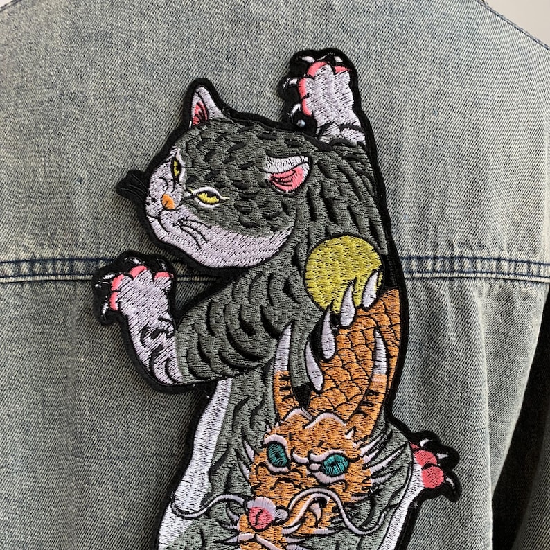 Peut inclure: Un &eacute;cusson brod&eacute; repr&eacute;sentant un chat gris aux yeux jaunes et aux pattes &agrave; coussinets roses tendant la main vers un orbe jaune, plac&eacute; au-dessus d'une t&ecirc;te de dragon orange aux yeux bleu sarcelle et aux longues moustaches sur du denim bleu clair.