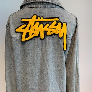 Puede incluir: Una chaqueta vaquera azul con un logotipo "STUSSY" bordado en amarillo en la espalda.