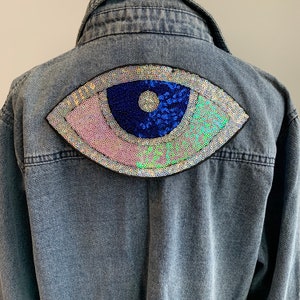 Może przedstawiać: Niebieska kurtka dżinsowa z dużym, tęczowym naszywką oka z cekinów na plecach.