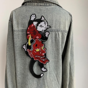Peut inclure: Une veste en jean gris avec un grand &eacute;cusson brod&eacute; au dos. L'&eacute;cusson repr&eacute;sente un chat noir et blanc plac&eacute; au-dessus d'un masque de d&eacute;mon japonais rouge avec des cornes jaunes, agr&eacute;ment&eacute; d'une grande fleur rouge et de d&eacute;tails noirs.