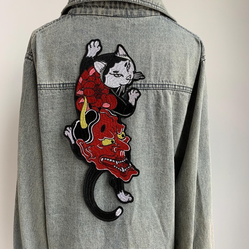 Peut inclure: Une veste en jean gris avec un grand &eacute;cusson brod&eacute; au dos. L'&eacute;cusson repr&eacute;sente un chat noir et blanc plac&eacute; au-dessus d'un masque de d&eacute;mon japonais rouge avec des cornes jaunes, agr&eacute;ment&eacute; d'une grande fleur rouge et de d&eacute;tails noirs.