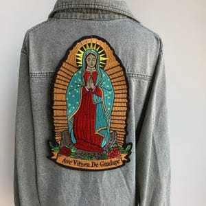 Op de afbeelding: Lichtgrijze spijkerjas met een grote geborduurde patch op de rug. De patch toont een religieuze figuur in een blauwe en rode jurk, omringd door een gouden halo. De tekst "Ave Virgen De Guadalupe" staat op een banner eronder.