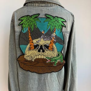 May include: Light blue denim jacket with a large embroidered patch on the back. The patch features a skull, palm trees, a skeleton, and an alligator, set against a tropical island backdrop.