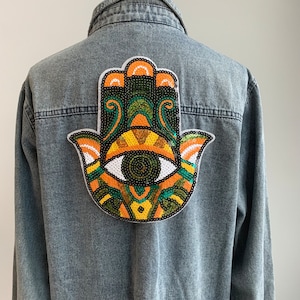 以下が含まれることがあります： 背中に大きなカラフルなハムサハンドパッチが付いた、明るいブルーのデニムジャケット。 パッチは、オレンジ、黄色、緑、黒のスパンコールでデザインされた目です。 ジャケットはクラシックな襟とボタン留めです。