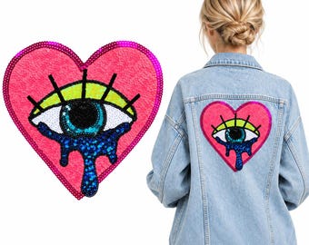 Op de afbeelding: Een lichtblauw spijkerjack met een grote hartvormige paillettenpatch op de rug. De patch toont een gestileerd oog met een geel ooglid, zwarte wimpers en een blauwe iris, met donkerblauwe pailletten die als tranen naar beneden lopen.