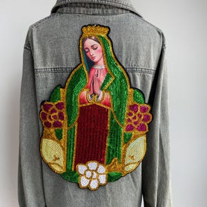 Puede incluir: Chaqueta vaquera gris con un parche grande y colorido en la espalda. El parche presenta una imagen de una figura religiosa con una corona dorada, ropa de lentejuelas verdes y rojas y un diseño floral. La chaqueta tiene botones y mangas largas.