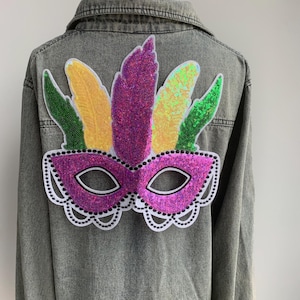 Könnte beinhalten: Graue Jeansjacke mit einem Pailletten-Mardi-Gras-Masken-Design auf dem Rücken. Die Maske ist lila mit weißen Verzierungen und Augenlöchern. Über der Maske befinden sich bunte Paillettenfedern in Gelb, Grün und Lila.